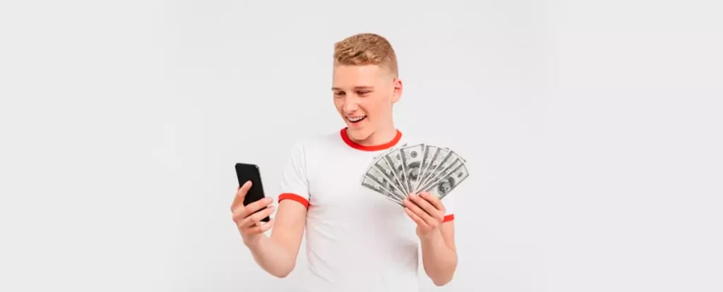 jovem adolescente feliz com celular na mão esquerda e dinheiro na mão direita em um fundo cinza claro