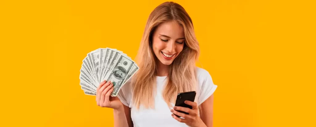 mulher com dinheiro na mão olhando para celular com fundo amarelo