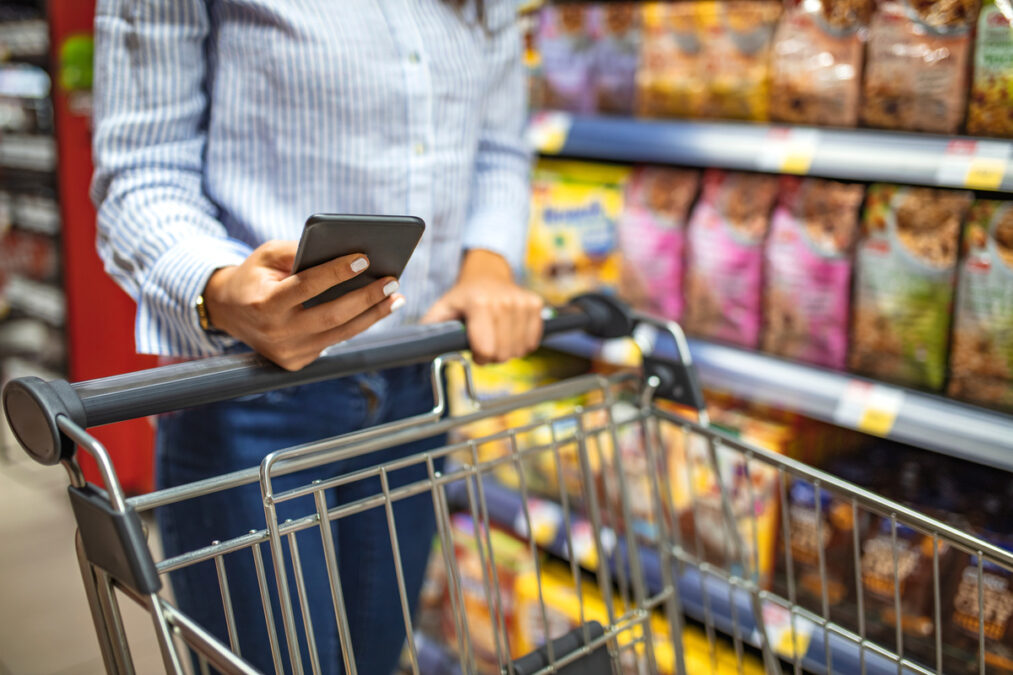 Imagem de mulher no supermercado olhando para celular e empurrando carrinho de compras