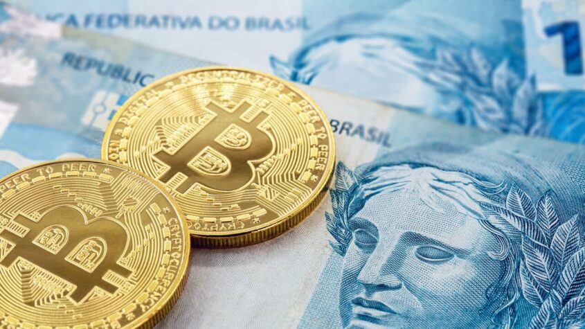 Imagem de notas de cem reais em baixo de moedas de Bitcoin
